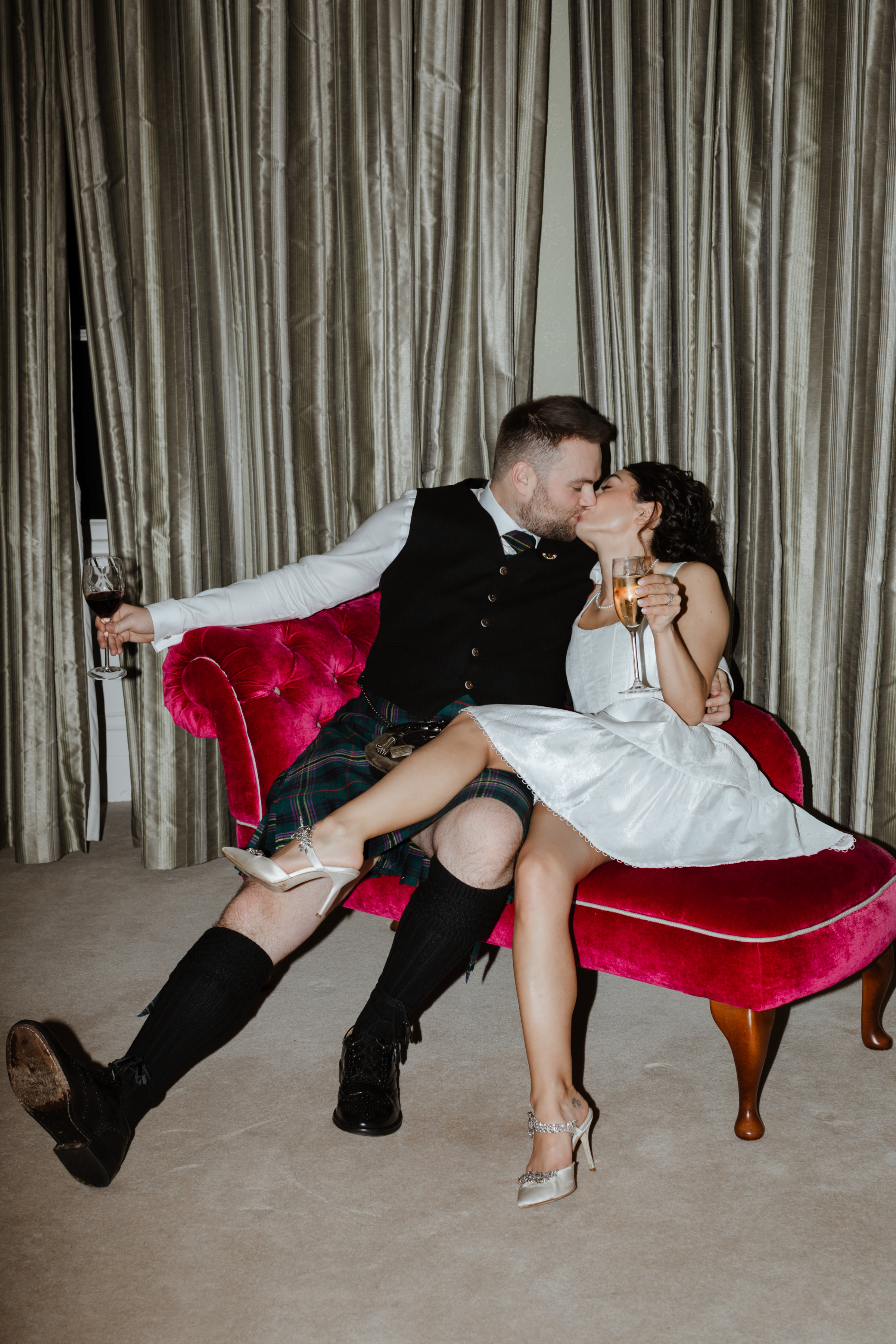 Bride in a short dress and groom in a kilt kissing on a red chaise longue. Caro Weiss Photography.